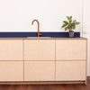 A modern kitchen cabinet with plyzen FSC certified birch plywood drawers and a dark blue countertop featuring a brass faucet, next to which sits a small potted plant with green and white leaves.